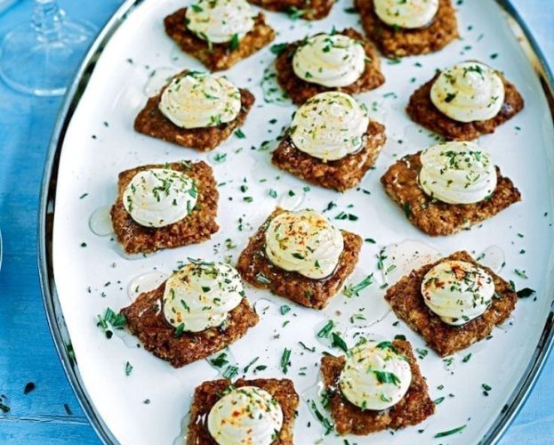 Lots of oat biscuits topped with goat's cheese and herbs shown on an oval plate