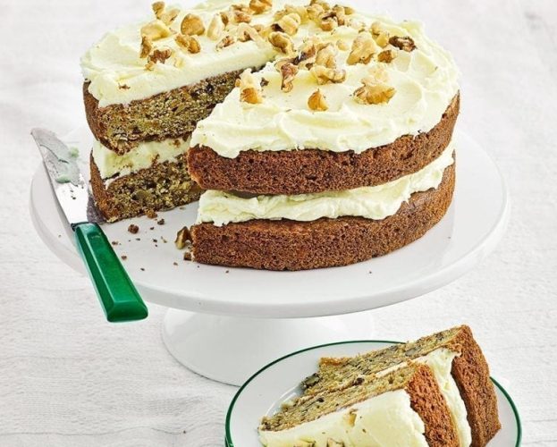 Iced carrot cake shown on a white cake stand, with a slice cut out