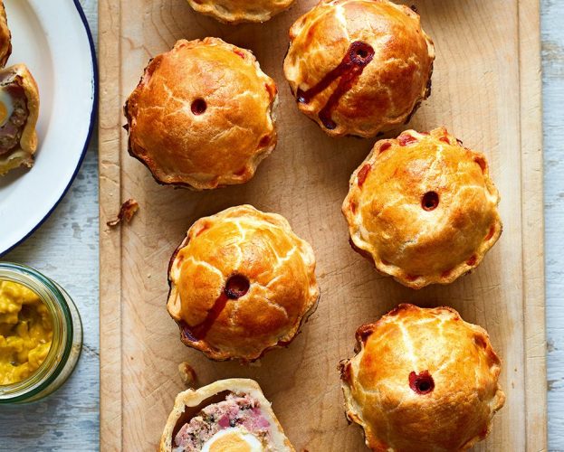 8 mini raised ham and egg pies sit on a wooden board, with one sliced open to reveal an egg