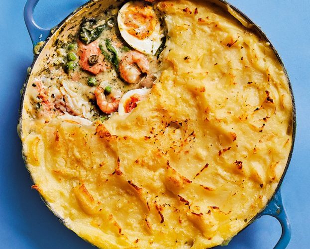 Fish pie with mashed potato topping in a blue casserole fish. A spoonful has been taken out, showing the filling, including a boiled egg and prawns