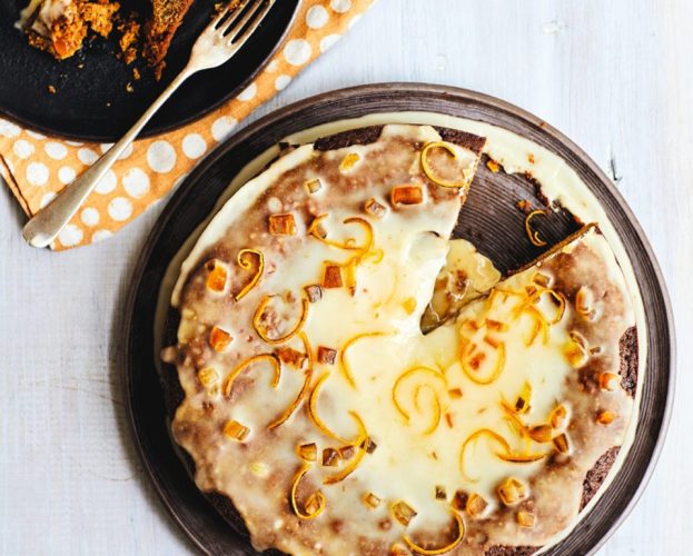 An aerial view of an iced marmalade cake, topped with chopped stem ginger and pared strips of orange zest, with a slice removed