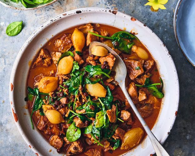 An overhead view of a casserole dish filled with pork stifado stew, topped with green spinach leaves