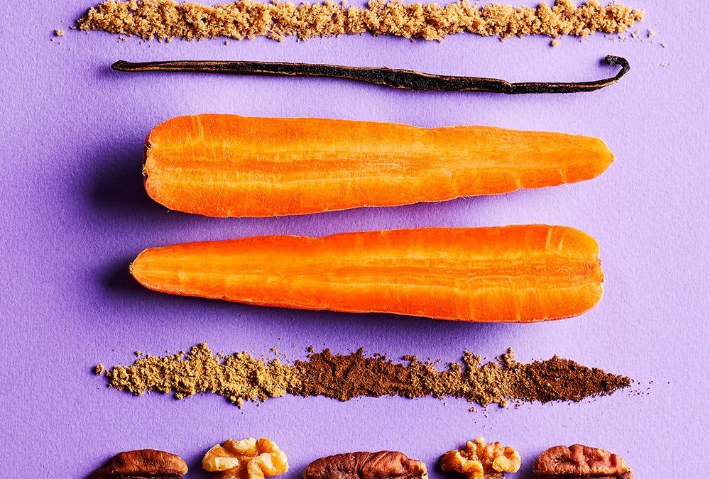The ingredients to make a carrot cake, including carrots, a vanilla pod, flour, sugar, sultanas and nuts, laid on a purple background
