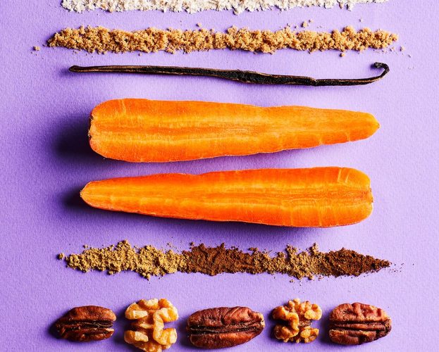 The ingredients to make a carrot cake, including carrots, a vanilla pod, flour, sugar, sultanas and nuts, laid on a purple background