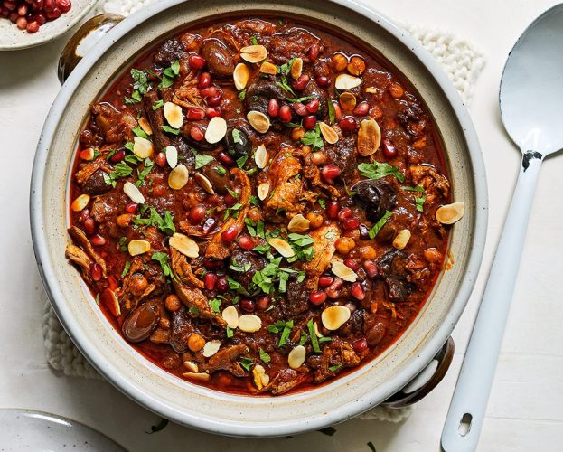 An overhead view of a pot of duck and prune tagine, topped with herbs, flaked almonds and pomegranate seeds