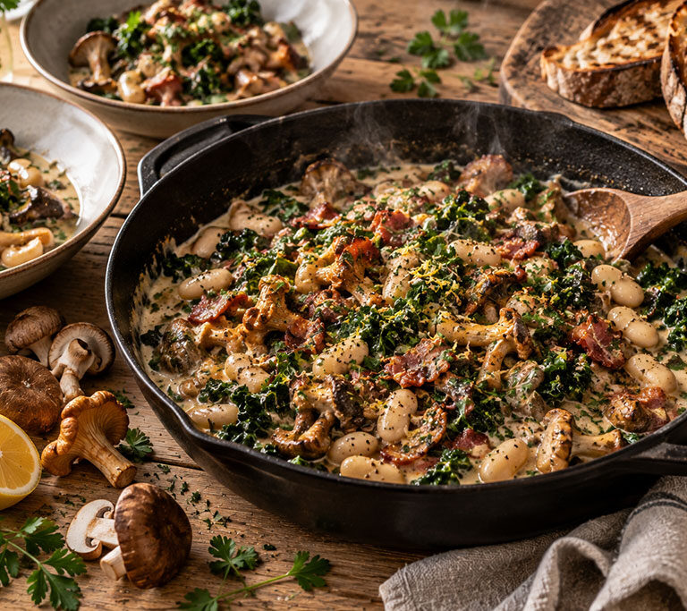 Cast-iron pan of wild mushroom and butter bean braise with kale, bacon and parsley, toasted sourdough alongside