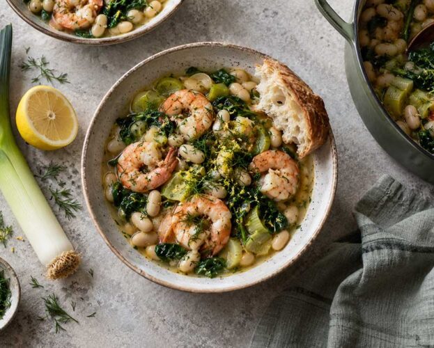 A bowl of creamy prawn, leek and white bean stew topped with dill, with crusty bread on the side