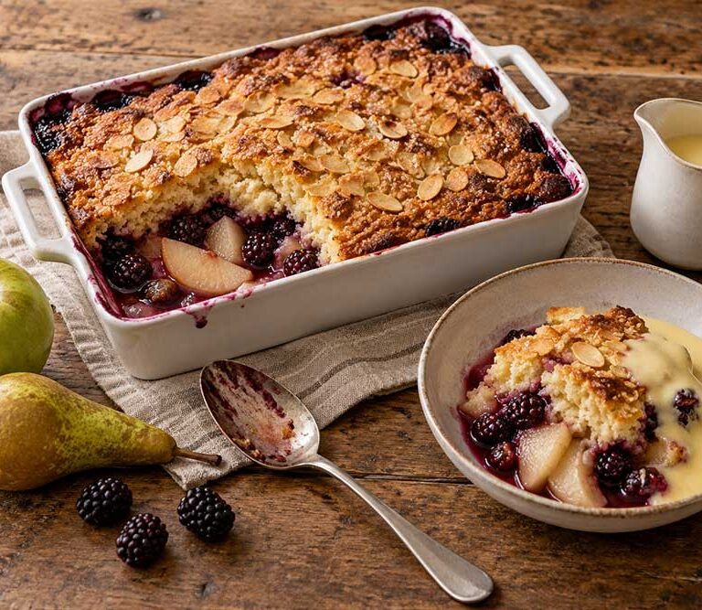 Apple, pear and blackberry orchard pudding in a baking dish with a golden almond sponge, one portion served with custard