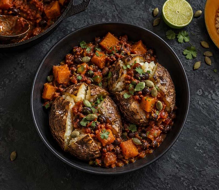 Pumpkin and black bean chilli jacket potatoes in a black bowl, topped with toasted pumpkin seeds and coriander